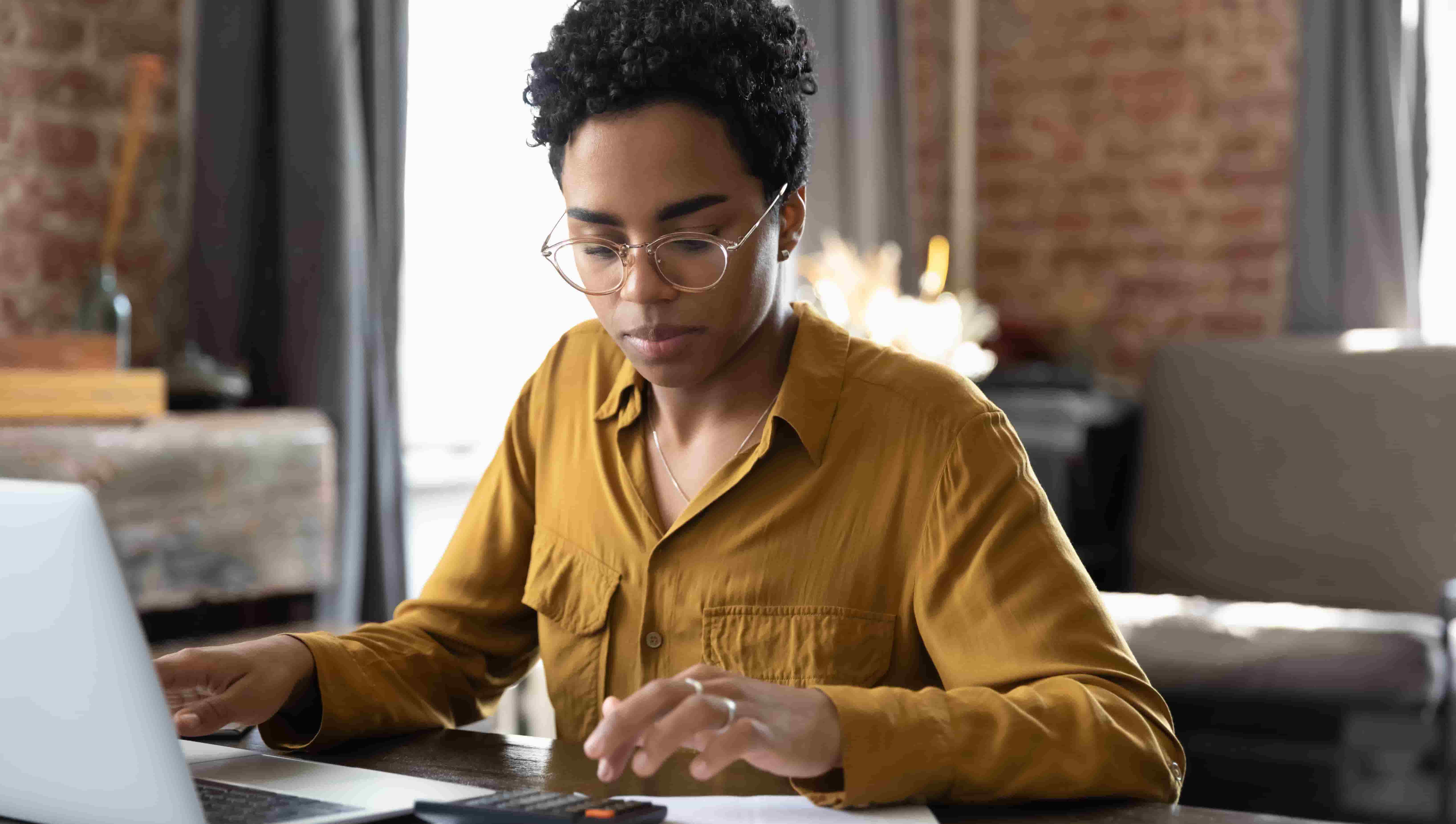 Focused millennial Black business woman calculating finance, money, using calculator, laptop computer at home workplace table, counting budget, paying bills, taxes, rent, mortgage fees